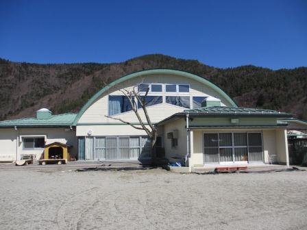 晴天と壮大な山をバックに建つ半月屋根の鳴沢保育所の写真